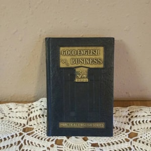 Può includere: Un libro blu vintage intitolato "Good English in Business" con lettere dorate e un emblema dorato. Il libro fa parte della "Practical English Series".