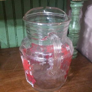 May include: A clear glass pitcher with a red and white floral design. The pitcher has a handle and a wide mouth.