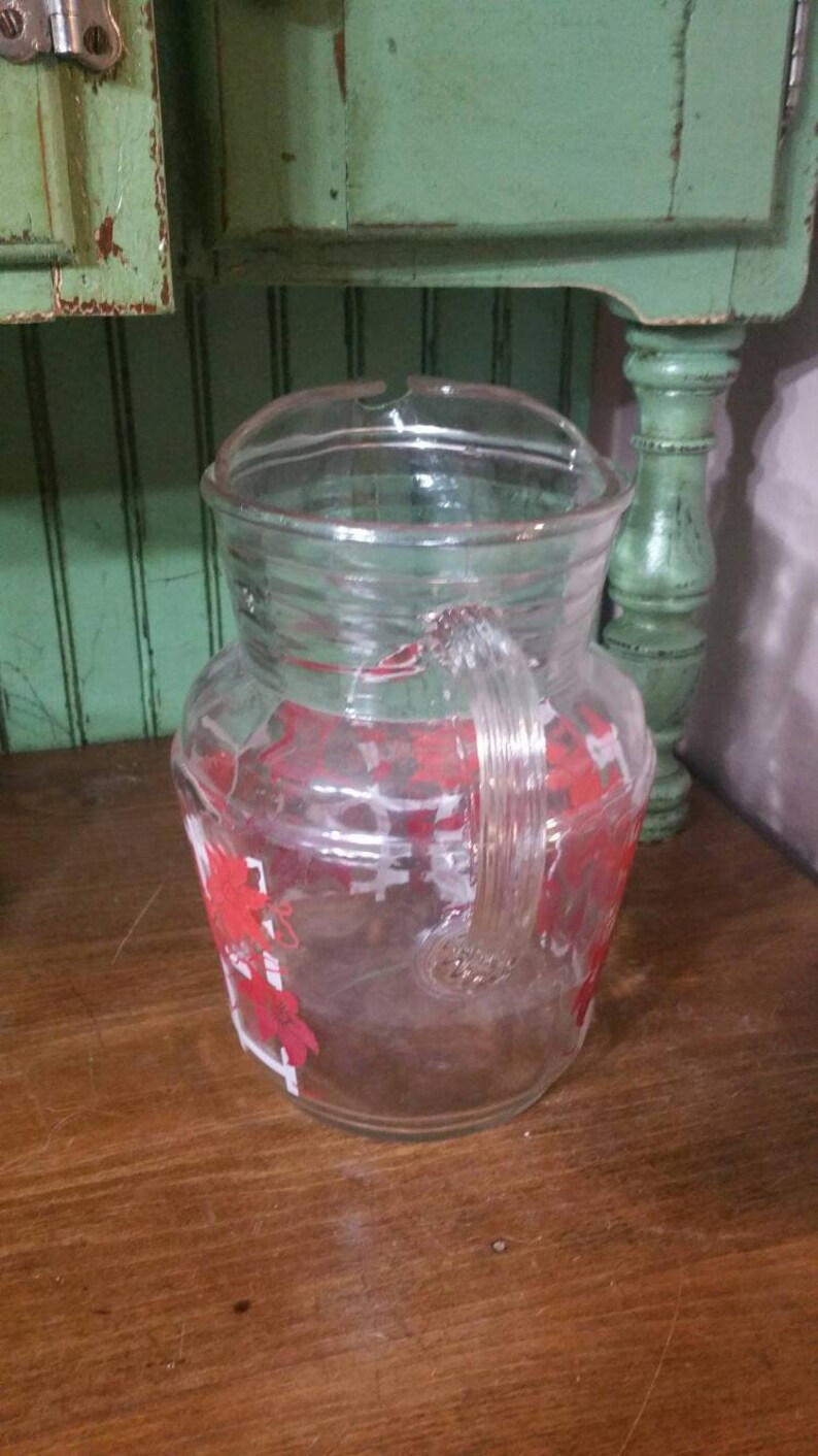May include: A clear glass pitcher with a red and white floral design. The pitcher has a handle and a wide mouth.