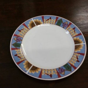 May include: A white ceramic plate with a blue, red, and yellow border featuring a pattern of sunflowers and houses.