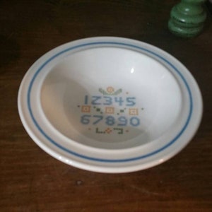 May include: White ceramic bowl with a blue rim and a blue and orange floral and number design in the center. The numbers 12345 and 67890 are displayed in a cross-stitch style.