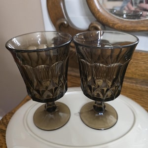 Puede incluir: Dos copas de vidrio vintage teñidas de marrón con detalles de patrón de diamantes en el cuenco. Las copas tienen una base con tallo y se exhiben en un plato ovalado blanco. El fondo incluye un espejo de madera.