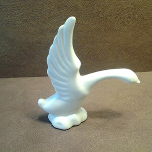 May include: A white ceramic goose figurine with outstretched wings. The goose is depicted in mid-flight, with its neck extended and head forward. The figurine is perched on a cloud-like base, creating a sense of movement and grace.