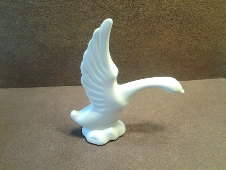 May include: A white ceramic goose figurine with outstretched wings. The goose is depicted in mid-flight, with its neck extended and head forward. The figurine is perched on a cloud-like base, creating a sense of movement and grace.