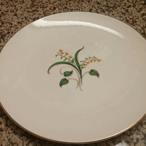 Könnte beinhalten: Ein weißer Keramikteller mit goldfarbenem Rand zeigt ein zartes Blumenmuster in Grün- und Gelbtönen. Das Muster zeigt eine stilisierte Blume mit Blättern und Stielen.