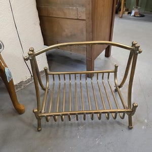 Puede incluir: Un revistero de latón vintage con una parte superior curvada y barras verticales. El revistero tiene una base rectangular y cuatro patas pequeñas. El latón tiene una pátina cálida y envejecida, lo que sugiere que es una pieza antigua o vintage.