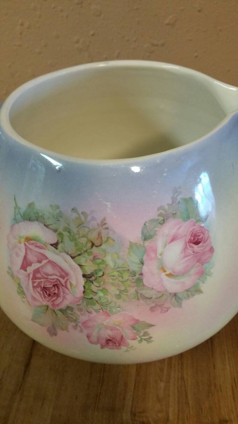 May include: A white ceramic jug with a blue and pink ombre glaze. The jug is decorated with a floral design of pink roses and green leaves.
