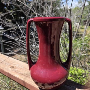 May include: A glossy, deep burgundy ceramic vase featuring a bulbous base, a slender neck, and two tall, symmetrical handles that extend from the shoulder to the rim. The surface has a smooth, reflective finish with a consistent dark red color.