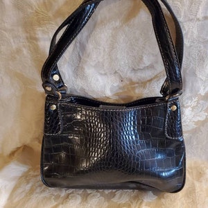 以下が含まれることがあります： クロコダイル柄のフェイクレザー製の黒いハンドバッグ。金色の金具が付いています。バッグにはショルダーストラップが2本付いています。