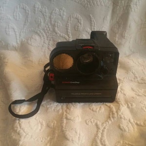 Peut inclure: Un appareil photo Polaroid OneStep noir avec un bouton rouge et un flash doré. L'appareil photo est doté d'une sangle noire et le texte "Polaroid ProntoLand Camera" est imprimé sur le devant.