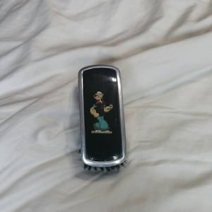 May include: Black and silver hairbrush featuring a cartoon image of Popeye the Sailor Man. The brush has a rectangular shape with a silver trim and black bristles. The Popeye image is in full color.