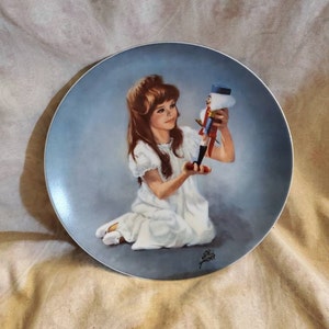 May include: A porcelain plate with a blue background depicts a painting of a young girl in a white dress holding a toy soldier. The girl has long brown hair and is looking at the soldier. The plate has a gold rim and a signature in the bottom right corner.