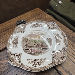 May include: A decorative, scalloped-edge plate with a brown and white transferware design. The central image depicts a castle scene with figures, trees, and water. The plate's border features floral and geometric patterns. The text "Belvoir Castle in 1793" is visible.