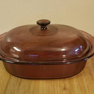 Può includere: Una pirofila in vetro vintage di colore viola scuro con coperchio. La pirofila ha un design a coste e una forma ovale arrotondata.