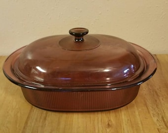 May include: A vintage, deep purple glass baking dish with a lid. The dish has a ribbed design and a rounded oval shape.