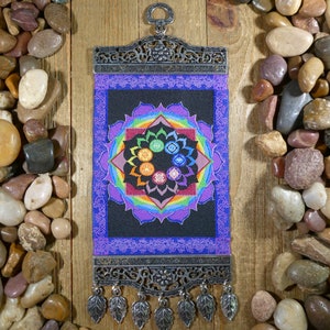 May include: A tapestry wall hanging with a black background and a colorful mandala design featuring seven chakras. The mandala is surrounded by a purple border and the tapestry is attached to a silver metal frame with a decorative design and a silver chain with leaf-shaped pendants.