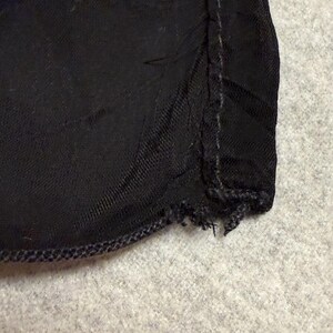 May include: Close-up of a section of black fabric. The fabric has a textured appearance and is stitched along the edges with a darker thread. The edge of the fabric is frayed in some areas.