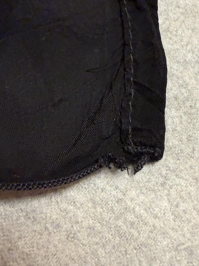 May include: Close-up of a section of black fabric. The fabric has a textured appearance and is stitched along the edges with a darker thread. The edge of the fabric is frayed in some areas.