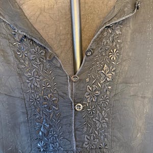 May include: Black blouse with floral embroidery around the neckline and button details. The blouse has a V-neck and a slightly textured fabric. The embroidery is in a darker shade of black, creating a subtle contrast.