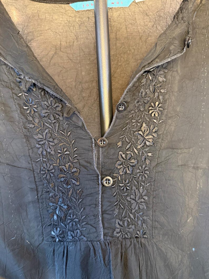 May include: Black blouse with floral embroidery around the neckline and button details. The blouse has a V-neck and a slightly textured fabric. The embroidery is in a darker shade of black, creating a subtle contrast.