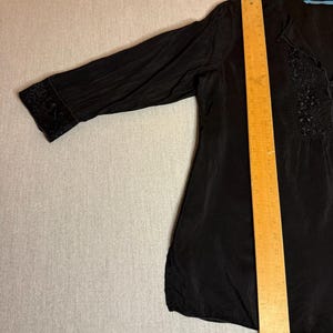 May include: Black blouse with long sleeves and a V-neck. The blouse has decorative embroidery on the cuffs and along the neckline. A wooden ruler is placed on top of the blouse.