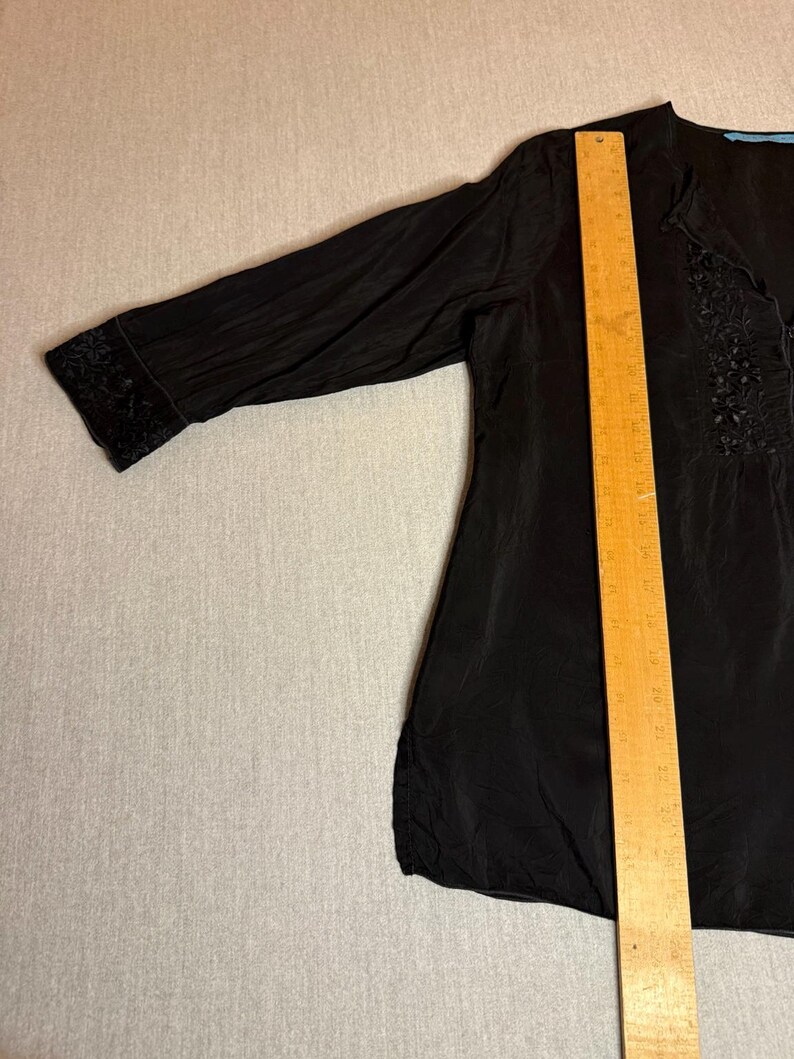 May include: Black blouse with long sleeves and a V-neck. The blouse has decorative embroidery on the cuffs and along the neckline. A wooden ruler is placed on top of the blouse.