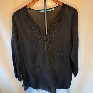 May include: Black long-sleeve blouse with a V-neck and button closure. The blouse features embroidered floral detailing around the neckline and cuffs. The blouse is hanging on a wooden hanger.