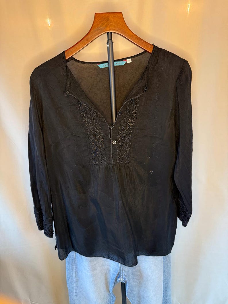 May include: Black long-sleeve blouse with a V-neck and button closure. The blouse features embroidered floral detailing around the neckline and cuffs. The blouse is hanging on a wooden hanger.