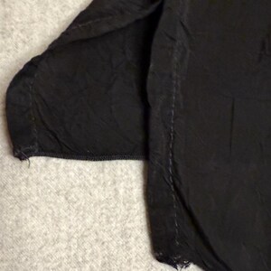 May include: Close-up of a black garment, possibly a shirt or blouse, with visible seams and a textured fabric. The edges show slight fraying, indicating a worn or vintage appearance. The fabric appears to be slightly wrinkled.
