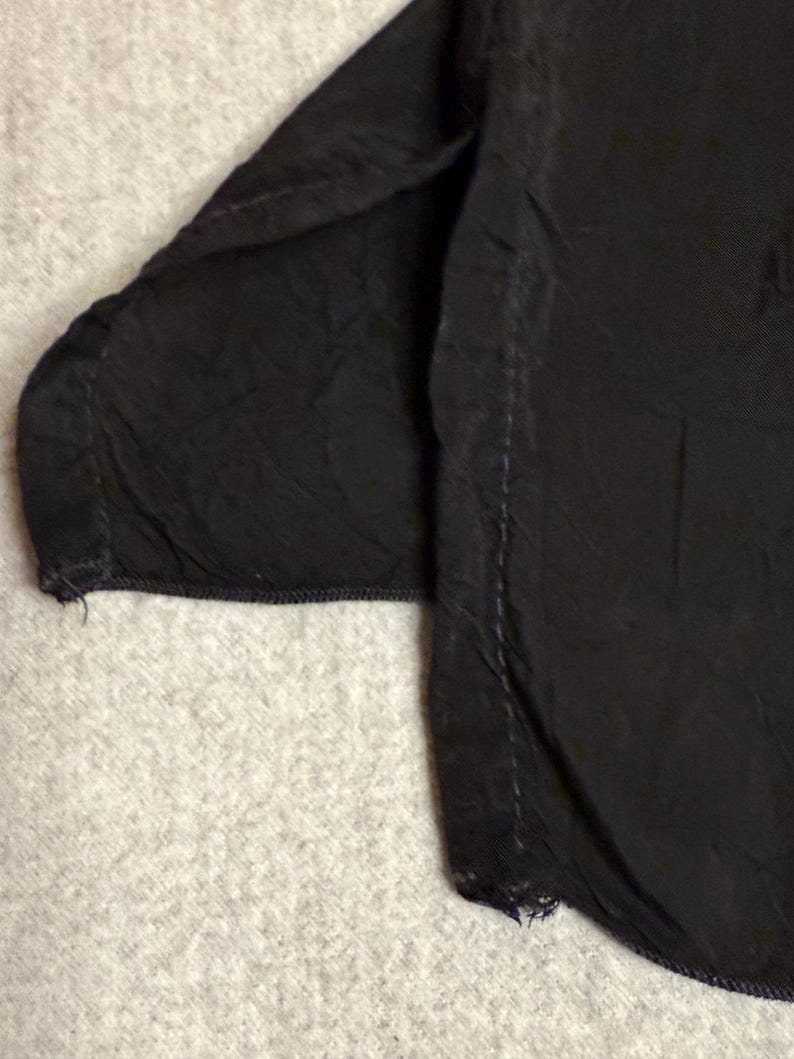 May include: Close-up of a black garment, possibly a shirt or blouse, with visible seams and a textured fabric. The edges show slight fraying, indicating a worn or vintage appearance. The fabric appears to be slightly wrinkled.