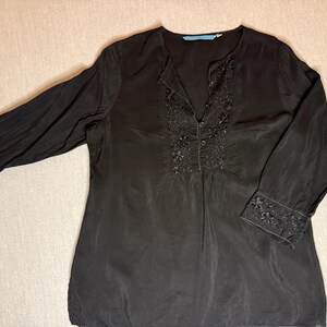 May include: Black long-sleeve blouse with floral embroidery around the neckline and cuffs. The blouse has a V-neck and a relaxed fit. The fabric appears to be lightweight and flowing, with a subtle sheen. The sleeves are long and end with embroidered cuffs.
