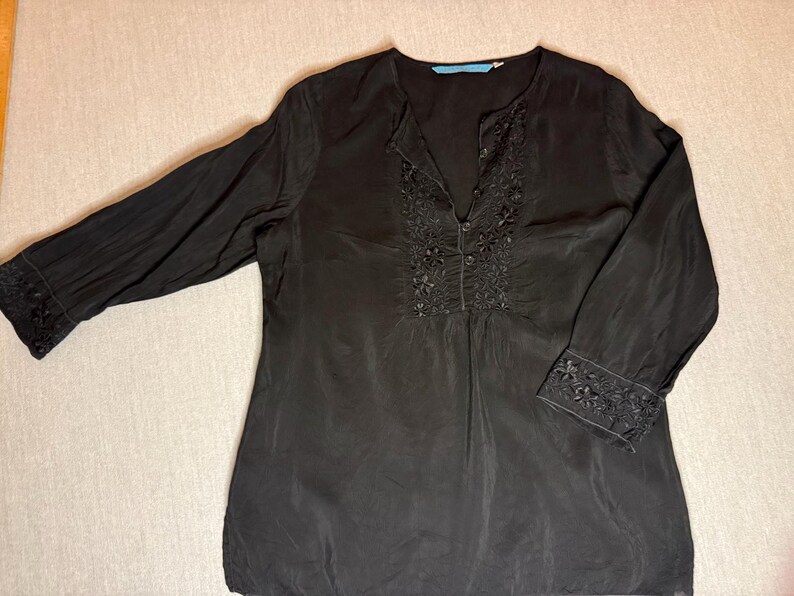 May include: Black long-sleeve blouse with floral embroidery around the neckline and cuffs. The blouse has a V-neck and a relaxed fit. The fabric appears to be lightweight and flowing, with a subtle sheen. The sleeves are long and end with embroidered cuffs.