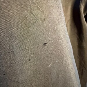 May include: Close-up of a textured, olive-green fabric. The fabric has a subtle sheen and visible weave, with small imperfections adding character. The image highlights the material's texture and color.