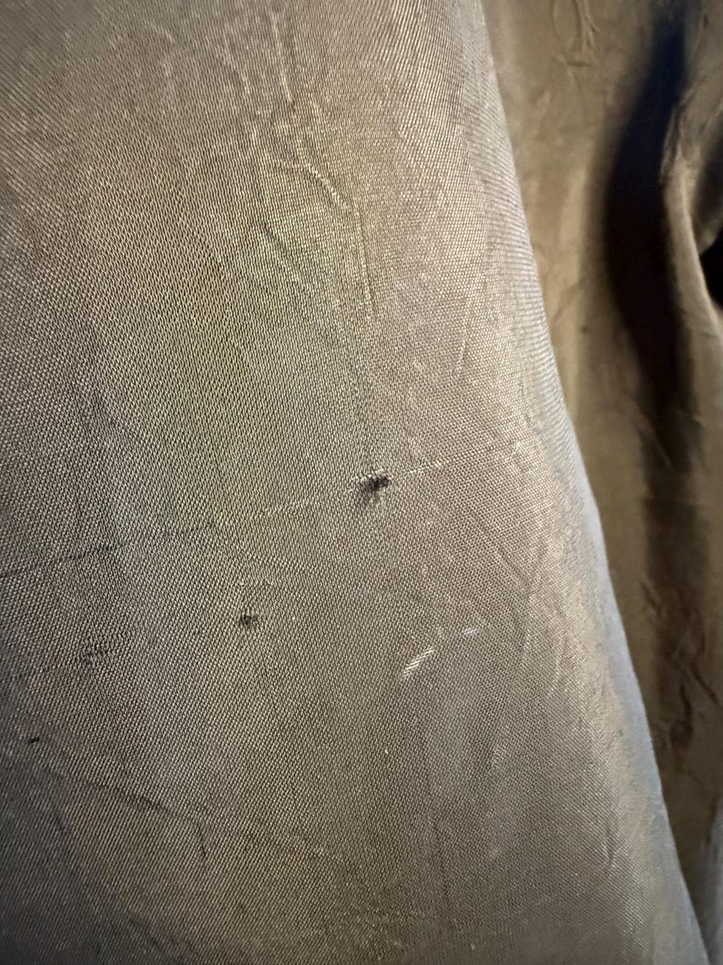 May include: Close-up of a textured, olive-green fabric. The fabric has a subtle sheen and visible weave, with small imperfections adding character. The image highlights the material's texture and color.