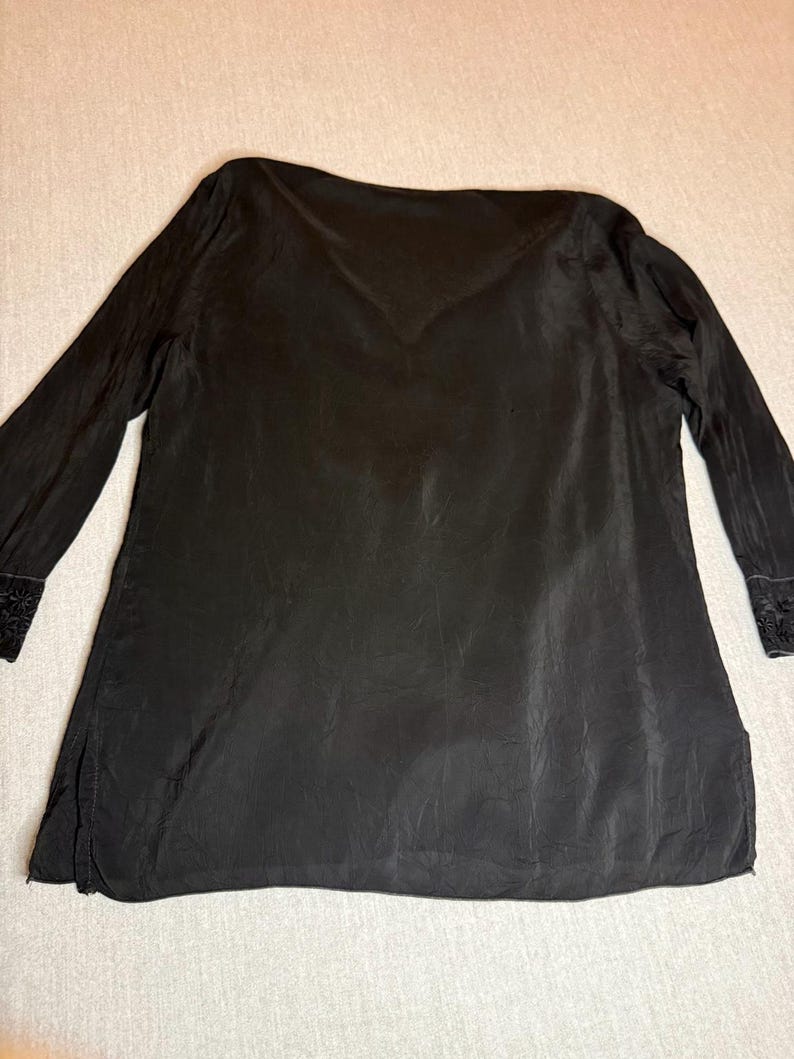 May include: Black long-sleeve tunic with a V-neck and a slightly textured appearance. The sleeves have decorative embroidery at the cuffs. The tunic is made of a lightweight fabric and has a loose, flowing fit.