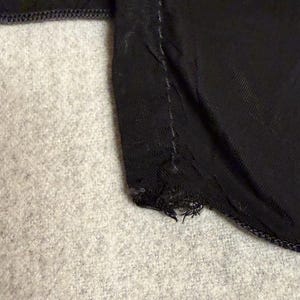 May include: Close-up of a black fabric with a visible seam and frayed edge. The fabric appears to be a dark, possibly satin-like material. The background is a light gray, textured surface.