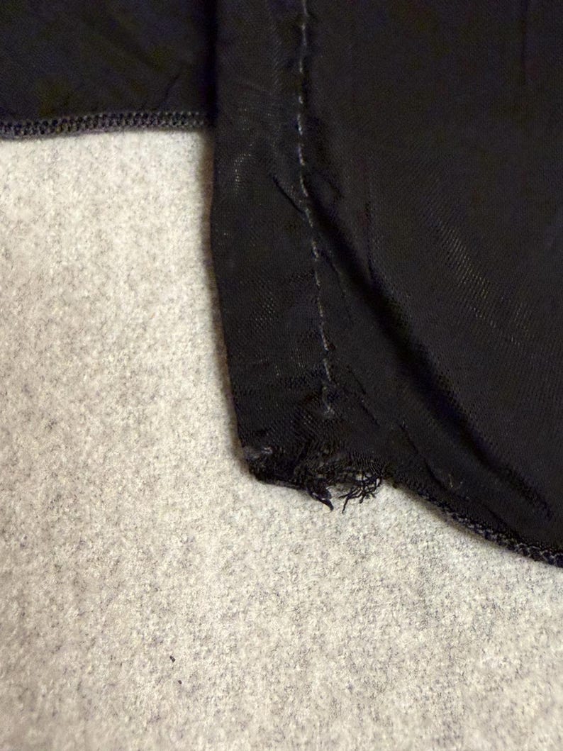 May include: Close-up of a black fabric with a visible seam and frayed edge. The fabric appears to be a dark, possibly satin-like material. The background is a light gray, textured surface.