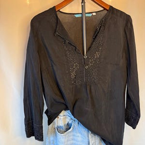 May include: Black long-sleeve blouse with floral embroidery on the front and cuffs. The blouse has a v-neck and button details. The garment is hanging on a wooden hanger, paired with light blue denim jeans.