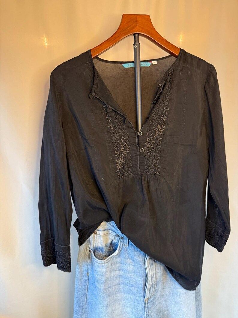 May include: Black long-sleeve blouse with floral embroidery on the front and cuffs. The blouse has a v-neck and button details. The garment is hanging on a wooden hanger, paired with light blue denim jeans.