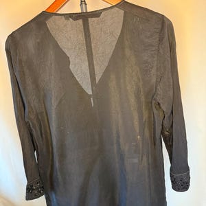 May include: Black long-sleeve blouse with a V-neck design. The blouse is made of a sheer, textured fabric and features decorative detailing on the cuffs. The garment is hanging on a wooden hanger.