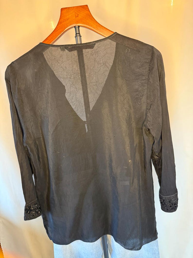 May include: Black long-sleeve blouse with a V-neck design. The blouse is made of a sheer, textured fabric and features decorative detailing on the cuffs. The garment is hanging on a wooden hanger.