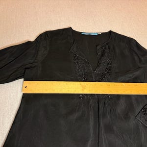 May include: Black long-sleeve blouse with floral embroidery around the neckline and cuffs. The blouse has a V-neck and a relaxed fit. A wooden ruler is placed across the blouse. The blouse measures approximately 20 inches across.