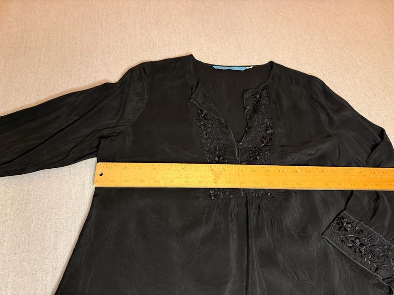 May include: Black long-sleeve blouse with floral embroidery around the neckline and cuffs. The blouse has a V-neck and a relaxed fit. A wooden ruler is placed across the blouse. The blouse measures approximately 20 inches across.