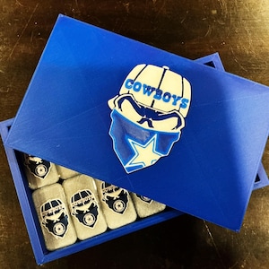 May include: A blue box with a lid featuring a Cowboys logo. Inside the box are several small, rectangular items with the same logo. The logo includes a cowboy hat, a bandana, and a star.
