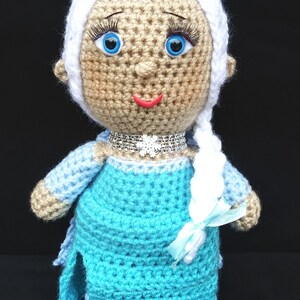 Pu&ograve; includere: Una bambola all'uncinetto con lunghi capelli bianchi, occhi azzurri e un vestito azzurro chiaro. La bambola indossa una collana argentata scintillante e un nastro azzurro chiaro.