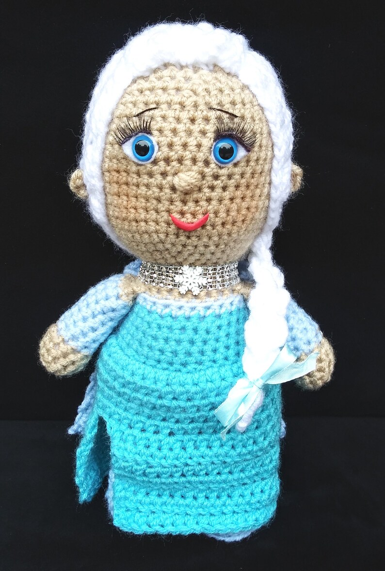 Pu&ograve; includere: Una bambola all'uncinetto con lunghi capelli bianchi, occhi azzurri e un vestito azzurro chiaro. La bambola indossa una collana argentata scintillante e un nastro azzurro chiaro.