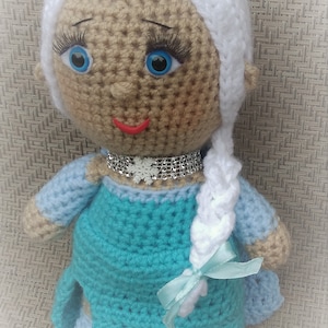 Pu&ograve; includere: Bambola all'uncinetto con una treccia bianca, un vestito blu e una collana con un fiocco di neve argentato. La bambola ha la pelle marrone, gli occhi blu e le labbra rosse.