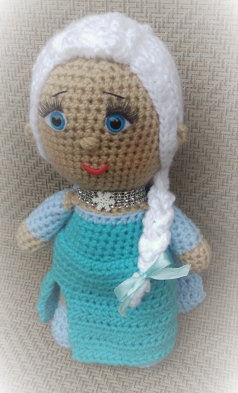 Pu&ograve; includere: Bambola all'uncinetto con una treccia bianca, un vestito blu e una collana con un fiocco di neve argentato. La bambola ha la pelle marrone, gli occhi blu e le labbra rosse.