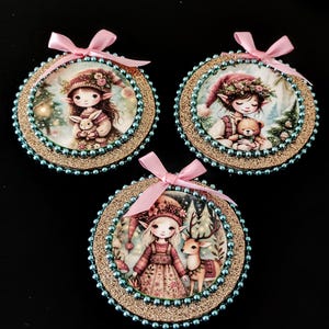 May include: Three round Christmas ornaments with illustrations of elves holding a bunny, teddy bear, and deer. Each ornament has a gold glitter border, blue bead trim, and a pink satin ribbon for hanging.
