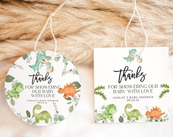 May include: Two round thank you tags with a dinosaur theme. The tags are white with green foliage and dinosaurs. The text on the tags reads "Thanks for showering our baby with love, Ashlee's Baby Shower, 09.24.22".